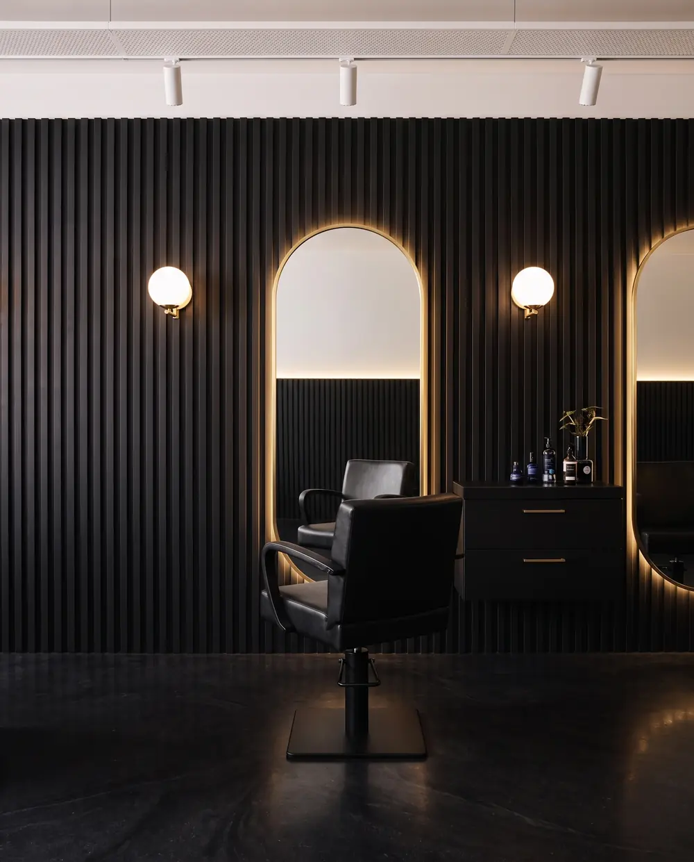 A black leather styling chair facing a tall gold arched mirror on a dark fluted wall